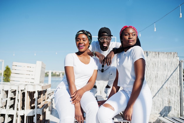 Trois amis afro-américains élégants portent des vêtements blancs à la jetée sur la plage Mode de rue des jeunes noirs Homme noir avec deux filles africaines