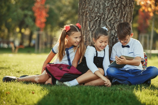 Trois amis d'école adolescentes souriantes assis dans le parc sur l'herbe et jouant ensemble nouveau jeu de tablette. Différentes émotions sur leurs visages