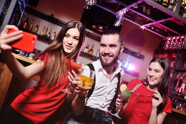 Trois amis, un jeune homme et deux jolies filles tenant des cocktails alcoolisés font un selfie de bar ou de boîte de nuit