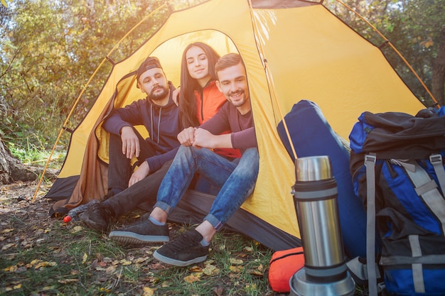 Trois amis sont assis dans une tente jaune et regardent dehors sur un sac à dos. Ils sourient un peu. Les gens sont en lieu sûr.