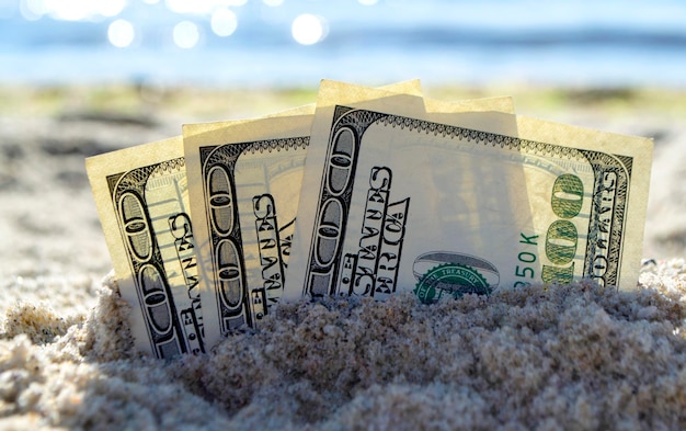 Trois billets d'un dollar sont enterrés dans le sable sur une plage de sable près de la mer le jour d'été ensoleillé en gros plan. Billets d'un dollar partiellement enterrés dans le sable