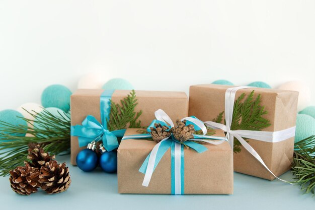 Trois boîtes de cadeaux de Noël enveloppées de papier artisanal rubans bleus et blancs décorés de branches de sapin cônes de pin et boules de Noël sur le fond bleu et blanc avec des lumières de Noël
