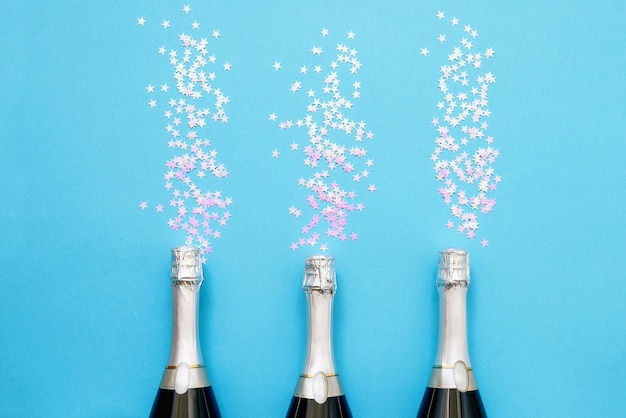 Trois bouteilles de champagne avec des étoiles de confettis holographiques sur fond bleu clair. Copiez l'espace, vue de dessus. Mise à plat de Noël, anniversaire, bachelorette, concept de célébration du nouvel an.