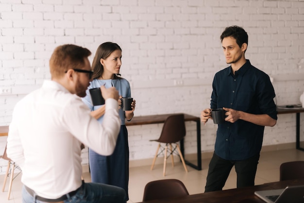 Trois collègues sympathiques debout au bureau, buvant du café et parlant joyeusement dans la salle de bureau pendant la pause sur fond de mur blanc en brique. Concept de la vie de bureau.