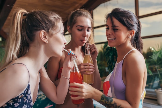 Trois copines heureuse en maillot de bain boire du jus de bouteilles à la piscine dans le centre de spa