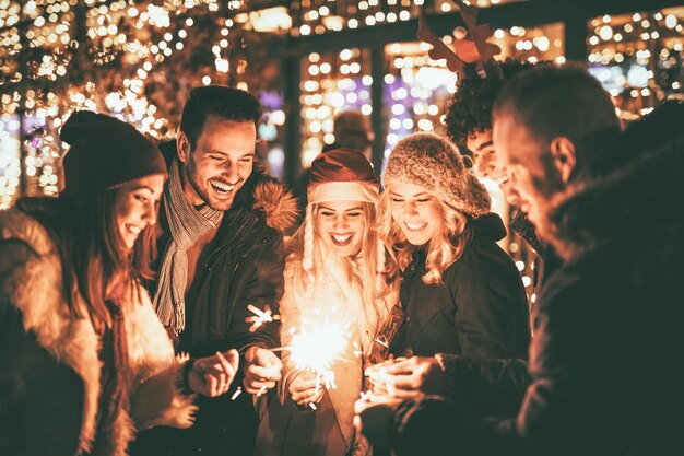 Trois couples avec des cierges magiques profitant de la fête de Noël en plein air dans la rue de la ville la nuit et avec beaucoup de lumières en arrière-plan.