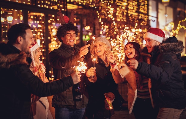 Trois couples avec des cierges magiques profitant de la fête de Noël en plein air dans la rue de la ville la nuit et avec beaucoup de lumières en arrière-plan.