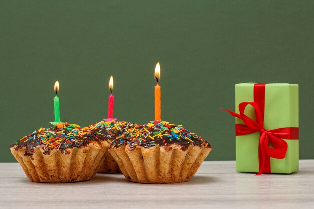 Trois délicieux cupcakes d'anniversaire avec glaçage au chocolat et caramel, décorés de bougies festives allumées et d'une boîte-cadeau sur fond vert. Concept minimal de joyeux anniversaire.