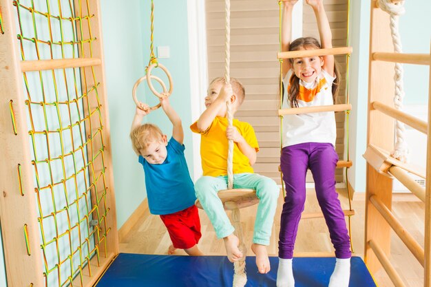 Trois enfants actifs d'âge différent grimpant sur l'échelle de corde en bois, la balançoire et les anneaux, jouant dans leur complexe sportif à domicile. Concept de mode de vie actif