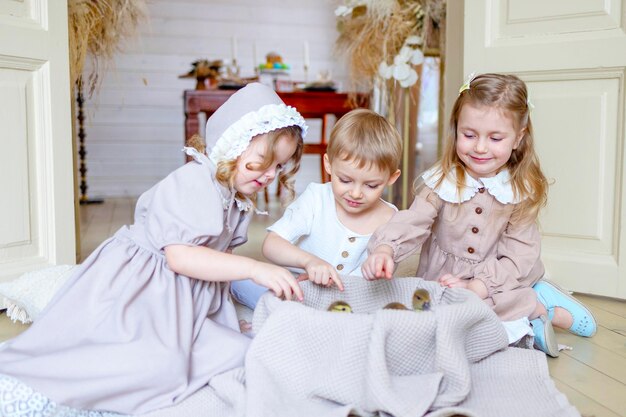 Photo trois enfants petites filles blondes en robe vintage et mignon petit garçon dans le salon maison de campagne le jour du printemps jouer avec les petits canards canards heureuse enfance simple vie confortable heureuse pâques