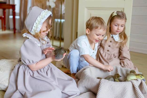 Photo trois enfants petites filles blondes en robe vintage et mignon petit garçon dans le salon maison de campagne le jour du printemps jouer avec les petits canards canards heureuse enfance simple vie confortable heureuse pâques