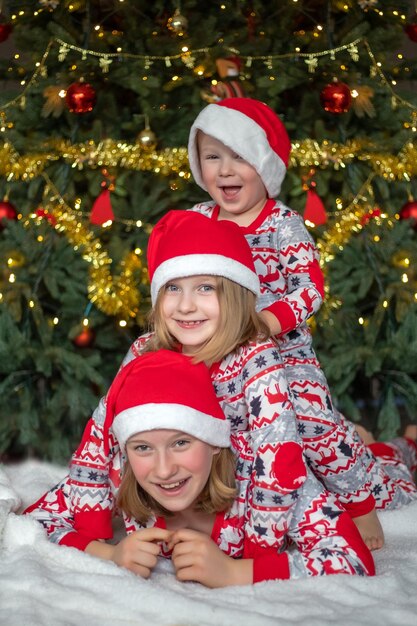 Trois enfants s'amusent sur le fond de l'arbre de Noël les enfants sont vêtus de pyjamas du Nouvel An et de chapeaux de père Noël