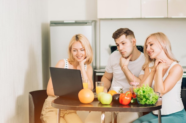 Trois étudiants en vêtements domestiques assis à table dans la cuisine, allant préparer le petit-déjeuner, regardant une recette vidéo sur un ordinateur portable, buvant du jus d'orange. Technologie, concept de personnes.