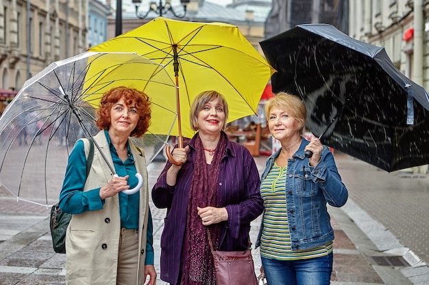 Trois Femmes âgées De Race Blanche Se Tiennent Sous Des Parapluies Dans La Rue De La Ville Européenne Par Temps Pluvieux.