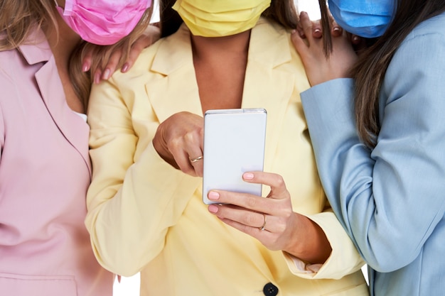 Trois femmes en costumes pastel posant avec des smartphones sur fond blanc