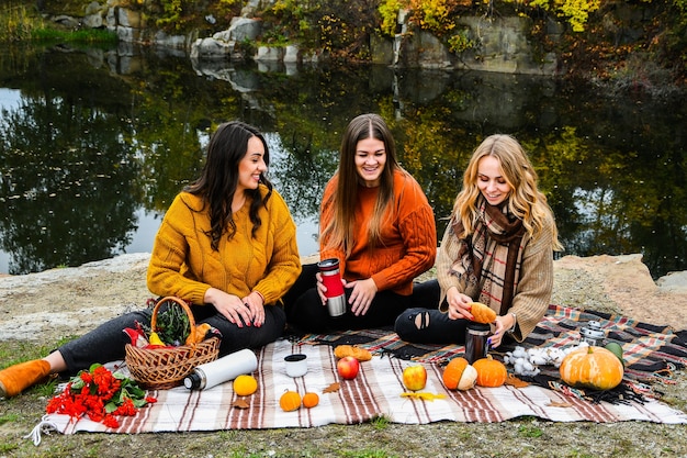 Trois femmes meilleures amies au pique-nique d'automne dans le parc. Plaid coloré, thermos et pompes. Amis s'amusant à l'extérieur. Chaude journée d'octobre d'automne