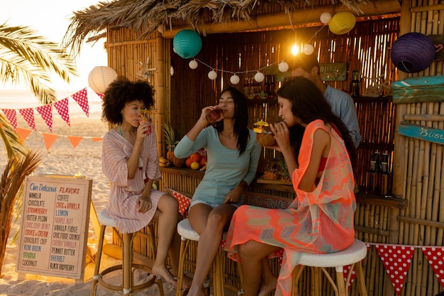 Trois femmes métisses profitant de leur temps à la plage avec leurs amis au coucher du soleil, assises près d'une cabane de cours de surf, buvant des boissons et souriant