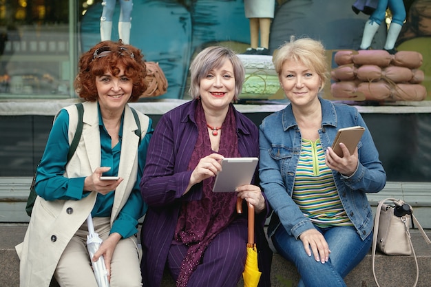 Trois femmes russes matures souriantes avec des appareils mobiles dans leurs mains sont assises sur la rue de la ville européenne en face de la fenêtre de l'épicerie.