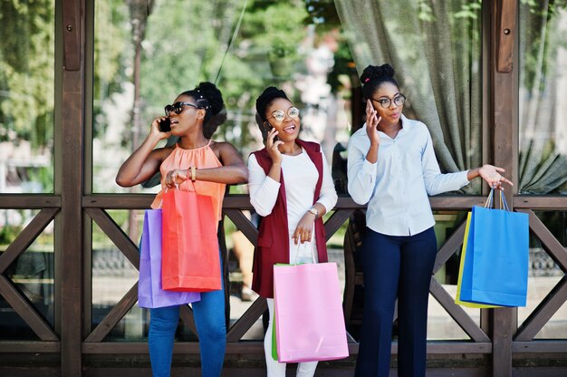 Trois filles afro-américaines décontractées avec des sacs à provisions colorés marchant en plein air. Femme noire élégante shopping et parlant sur téléphone mobile.