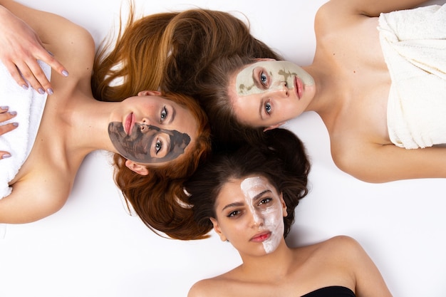 Trois filles, copines avec des demi-masques différents, sont allongées sur le sol, enveloppées dans des serviettes. Peau saine et concept de beauté sur un mur blanc. Photo de haute qualité