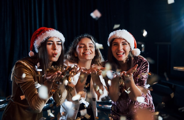 Trois filles positives soufflant des confettis dorés de leurs mains.