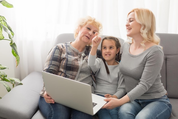 Trois générations de femmes heureuses s'assoient se détendent sur un canapé en riant en regardant une vidéo amusante sur un ordinateur portable, des femmes souriantes et positives, la grand-mère, la mère et la fille s'amusent se reposent sur le canapé