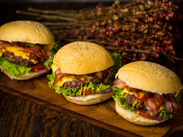 Trois hamburgers avec escalope de burger de boeuf, oignon frit, épinards, sauce ketchup et fromage, servis sur une planche à découper en bois sur fond de bois foncé.