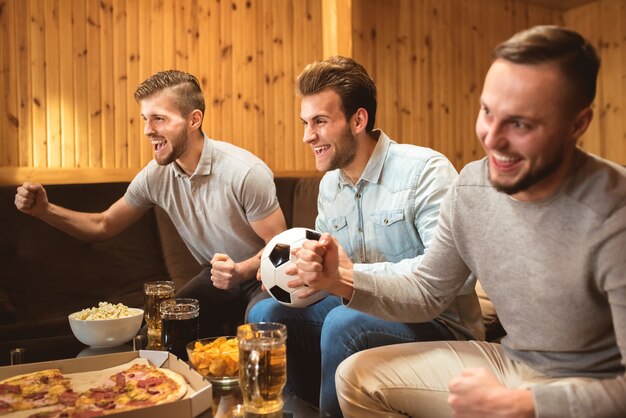 Les trois hommes avec une bière et une nourriture regardent un football et font un geste