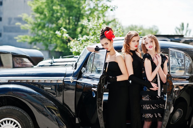 Trois jeune fille en robe de style rétro près de vieilles voitures anciennes classiques.