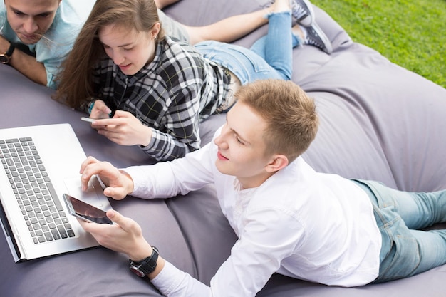 Trois jeunes amis allongés sur un grand coussin et se détendant à l'extérieur avec leurs téléphones et un ordinateur portable. Concept d'éducation ou d'amitié.