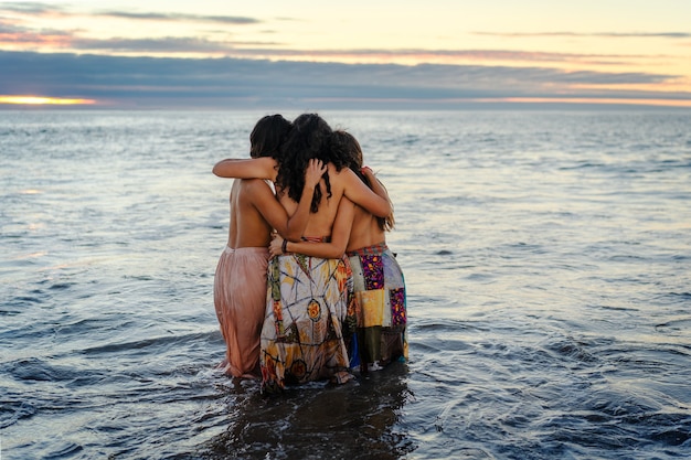 Trois jeunes femmes latines s'embrassent dans l'eau au coucher du soleil montrant l'amitié ou la fraternité