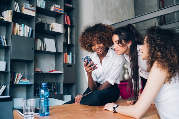 Trois jeunes femmes multiraciales à la recherche d&#39;un smartphone dans un bureau moderne co-working