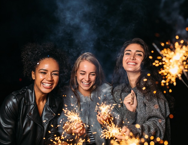 Trois jeunes femmes rient la nuit et tiennent des étincelles.