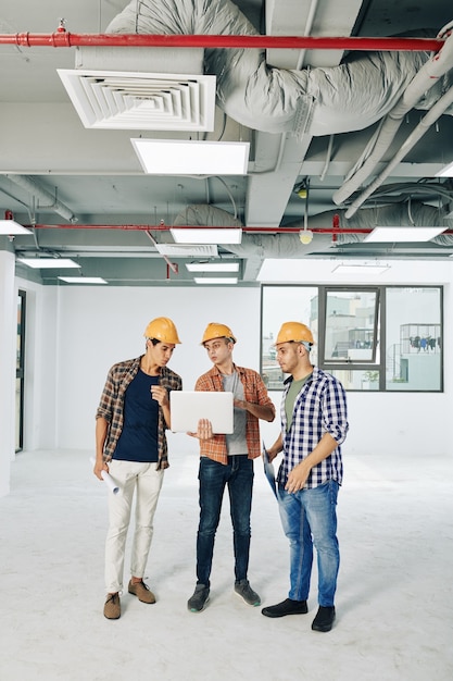 Trois jeunes ingénieurs civils discutant du schéma des lignes de service sur l'écran d'un ordinateur portable lorsqu'ils travaillent dans un bâtiment en construction