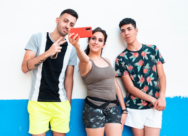 Trois jeunes, en vêtements d'été, sur un mur blanc et bleu, à l'aide de leurs smartphones