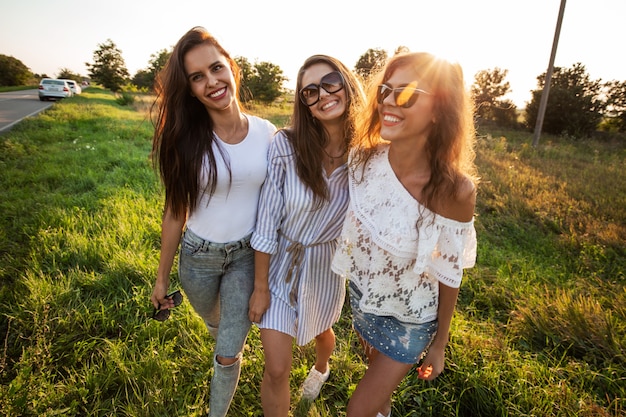 Trois magnifiques jeunes femmes à lunettes de soleil vêtues de beaux vêtements se tiennent sur le terrain et sourient par une journée ensoleillée. .