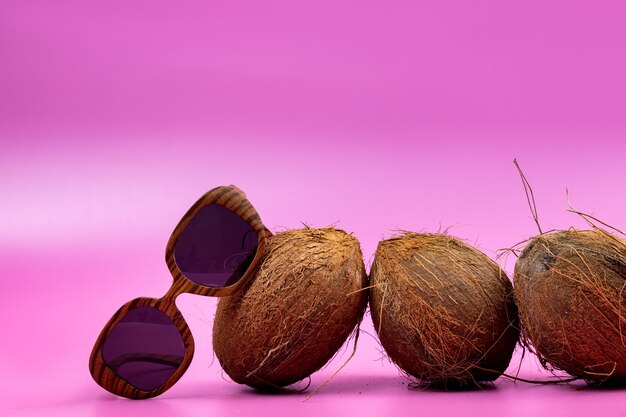 Trois noix de coco entières et verres en bois sur fond rose.