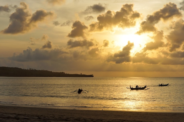 Trois pirogues se découpant au crépuscule sur la surface rougeoyante de la mer sous un coucher de soleil orange