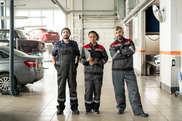 Trois professionnels interculturels sérieux en vêtements de travail debout devant la caméra dans un grand atelier à la fin de la journée de travail