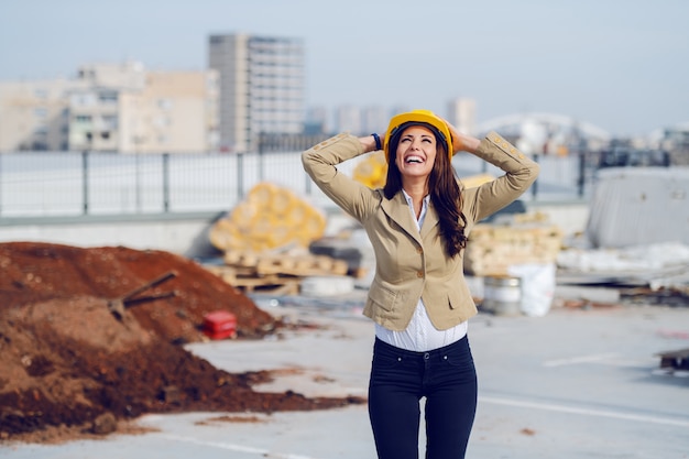 Trois quarts de longueur de l'architecte femme caucasienne heureuse en veste beige et avec un casque de protection sur la tête et en levant en se tenant debout sur le chantier de construction.