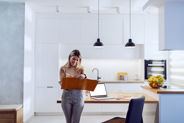 Trois quarts de longueur de femme d'affaires jolie blonde caucasienne en pull s'appuyant sur la table de la cuisine et d'écrire des statistiques. Sur la table de la cuisine, il y a un ordinateur portable et de la paperasse.