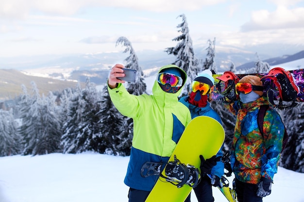 Photo trois snowboarders prenant un selfie avec l'appareil photo d'un smartphone dans une station de ski amis photographiant pour le partage sur les réseaux sociaux avec des snowboards près de la forêt portant des lunettes réfléchissantes vêtements de mode colorés