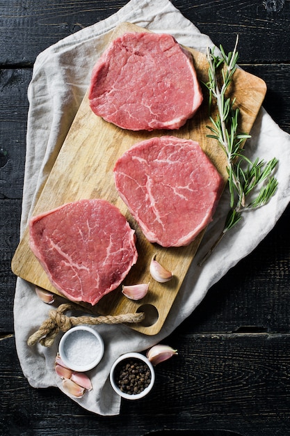 Trois steaks de bœuf crus sur une planche à découper en bois, de l&#39;ail et un brin de romarin.