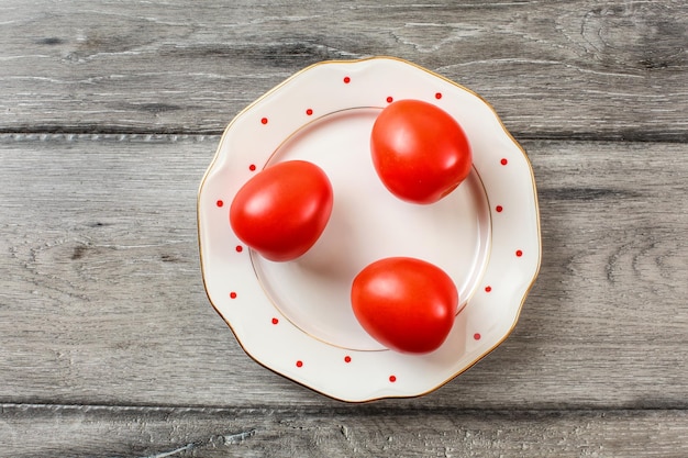 Trois tomates entières sur une assiette blanche avec de petits points rouges et un bord doré, un bureau en bois gris, une vue sur la table.