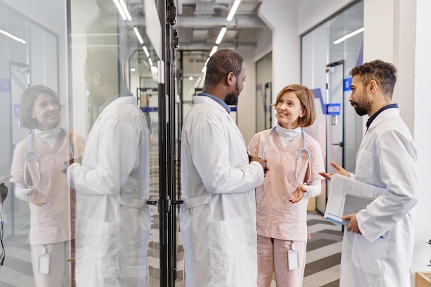 Trois travailleurs médicaux professionnels contemporains en blouse de laboratoire discutant lors d'une réunion de travail dans le couloir des cliniques