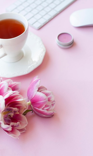 Trois tulipes de pivoine roses et tasse de thé noir, clavier et souris sans fil blancs, baume à lèvres sur fond rose