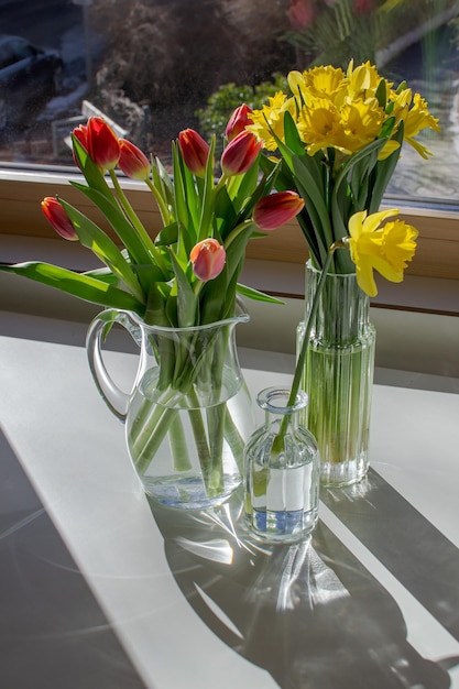 Trois vases en verre avec des tulipes et des jonquilles sur une table blanche au soleil