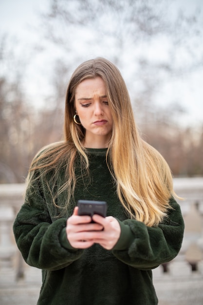 Troublée jeune femme se frottant le front et regardant un smartphone moderne tout en se tenant dans le parc en automne