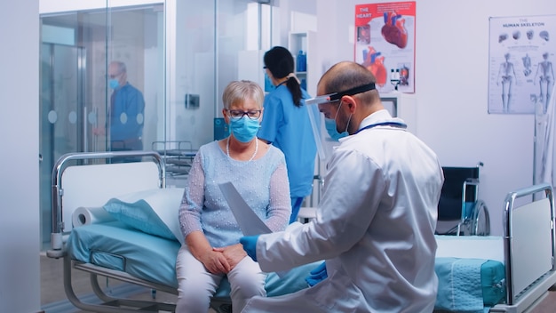 Des trucs médicaux dans la visière expliquant le résultat X Ray à une vieille femme à la retraite assise sur un lit d'hôpital pendant la pandémie de COVID-19. Infirmière vérifiant avec le vieil homme en arrière-plan. Clinique privée moderne ou faci de soins de santé