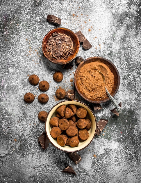 Truffes au chocolat, poudre de cacao et chocolat râpé dans des bols. Sur une table rustique.